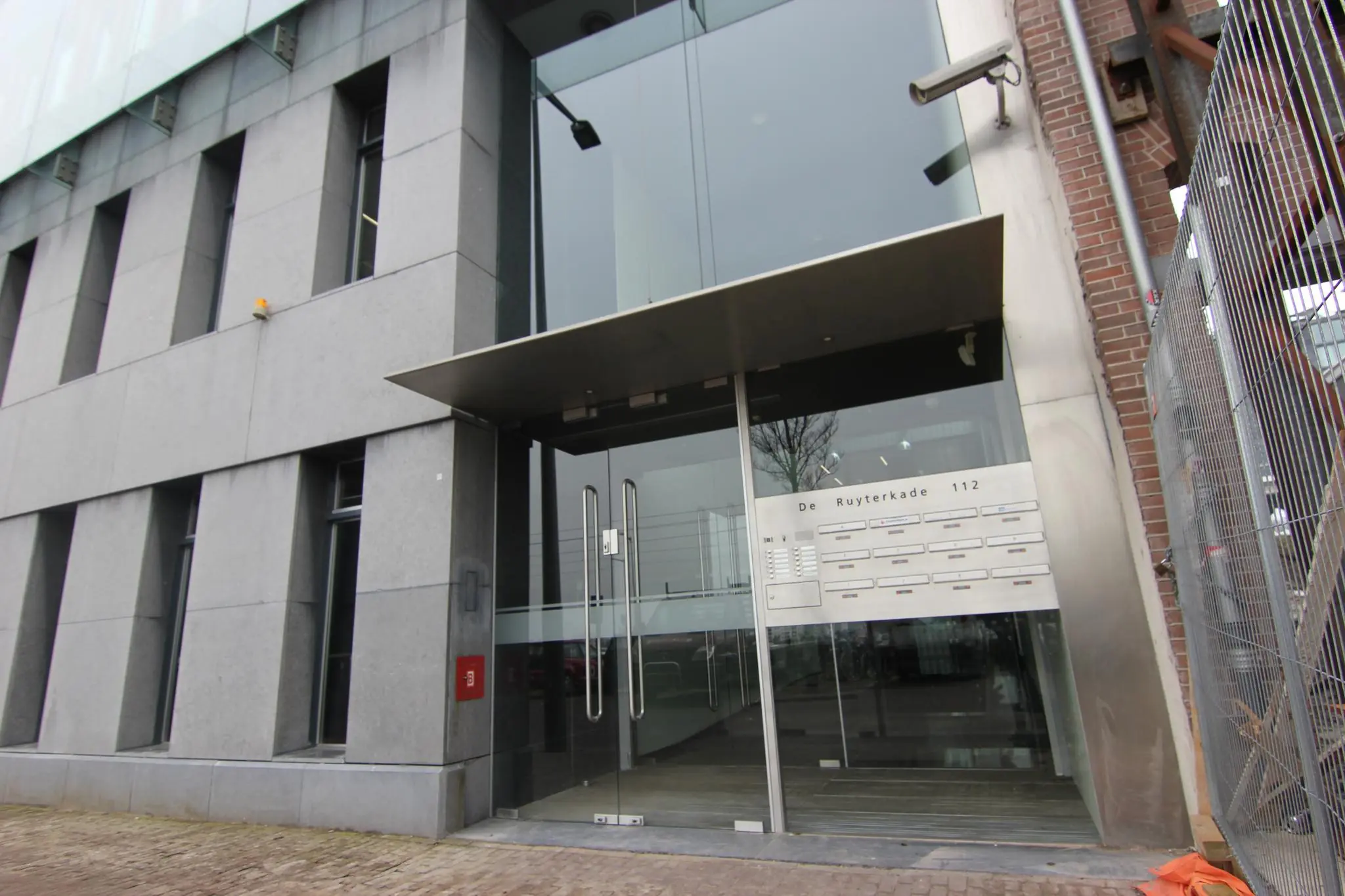 Ingang van een modern kantoorgebouw aan de De Ruyterkade 112 met glazen deuren en een intercompaneel.